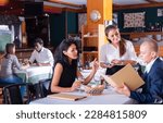 Small photo of positive female busboy taking order to couple in modern restaurante