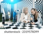 Small photo of Senior woman and her granddaughter thinking about chess riddle solution in escape room. Old man and grandson resolving other riddles in background.