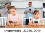 Small photo of Two teenage girls sitting at table in kitchen, engrossed in smartphones, oblivious to surroundings. Parents standing behind in blurred background also occupied with mobile gadgets