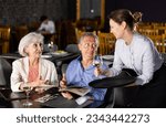 Small photo of Amiable female owner of restaurant bringing glasses of wine to positive senior couple, sitting at table and choosing dishes from menu card