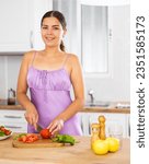 Small photo of Portrait of positive girl in nightdress standing at kitchen table at home, slicing vegetables, cooking salad.
