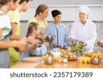 Small photo of Amiable woman, qualified chef wearing white cook jacket and toque, running culinary courses for preteen children, sharing secrets of cooking ..