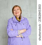 Small photo of Amiable smiling senior woman with ginger hair wearing light purple jacket posing with crossed arms on gray studio background. Positivity and confidence emanating from female countenance and stance