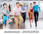Small photo of Cheerful adult man participating corporate party in light spacious office hall on Christmas Eve, dancing energetic upbeat jive with young brunette in blue dress with silver tinsel around neck