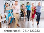 Small photo of Cheerful adult man participating corporate party in light spacious office hall on Christmas Eve, dancing energetic upbeat jive with young brunette in blue dress with silver tinsel around neck
