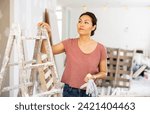 Small photo of Thoughtful asian woman standing at stepladder in apartment during repair works.