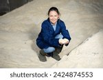 Small photo of Positive woman agronomist squatting at bunch of corn flour inside of fodder storage.