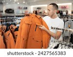 Small photo of After inspection of stores assortment, guy chooses weather-resistant jacket on hangers and closely examines its details, feels texture of fabric. Buyer goes through folds touch of overcoat fabric