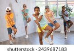 Small photo of African girlie perform movements during warm-up, limbering-up part of workout with peers. Group of young girls and guys in ball cap and dark glasses dance in fitness club in background unfocused