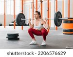 Small photo of Full body female athlete in activewear looking away and doing barbell back squat during intense workout in sunlit gym
