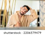 Reflection of young male in soft bathrobe touching dark hair and looking in mirror during spa session in salon