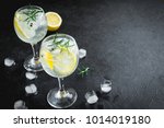 Alcohol drink (gin tonic cocktail) with lemon, rosemary and ice on rustic black stone table, copy space, top view. Iced drink with lemon and herbs.