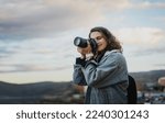 Small photo of Young cheerful woman traveler in a denim jacket with a backpack holds a camera in her hands while standing on the shore of the mountain lake