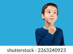 Small photo of Pensive kid looks away at copy space thinking isolated on a blue background, funny kid lips hold finger near mouth conceiving some kind of joke, conceptual image