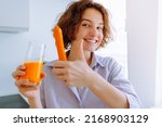 Small photo of young woman smiling showing thumbs up sign to super carrot fresh in home kitchen. teenage who drinks vitamin drink in morning from freshly squeezed carrot juice enriched with carotene and vitamin A
