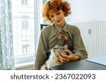 Small photo of Portrait attractive teenage girl with red hair, wearing hipster clothes, shirt and cravat, holding fluffy domestic cat in arms, purring. Home life pets and their owners, real life