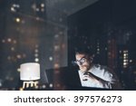 Bearded young businessman working on modern loft office at night. Man using contemporary notebook texting message, holding cup espresso, blurred background. Horizontal, film effect, bokeh