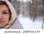 A young woman stands against a background of fir trees under a snowy forest in winter. A girl in a gray down jacket with a hood stands and looks at the camera. Close-up.