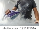 The hot steam from the iron. Powerful film effect of steam on photography. A close-up of a man's body in a grey t-shirt ironing clothes on an ironing board