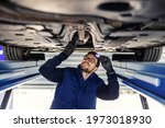 Technical inspection of the car. Car service in the workshop. A man in a blue uniform stands under a car in the garage and checks the car’s axles. It illuminates the chassis with a flashlight