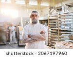 Small photo of A successful food factory supervisor in a white sterile uniform is standing in the factory with arms crossed. He is very satisfied with the quality and quantity of products.