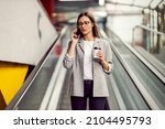 Small photo of A busy young businesswoman dressed elegantly descending the escalator while holding takeaway coffee and talking on the phone with a client. A businesswoman on the escalator with phone