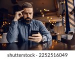 Small photo of A worried man is sitting in cafeteria and looking at his phone.