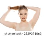 Front view of a young blond woman tying her long hair on a white studio background
