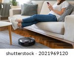 Robot vacuum cleaner cleaning the living room. Young woman enjoy rest, sitting on sofa at home