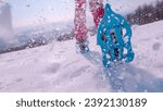 Small photo of LOW ANGLE VIEW, CLOSE UP: Active lady running in blue snowshoes and cloud of snow is flying behind her and sparkles in winter sunlight. Following unknown woman snowshoeing through snowy countryside.