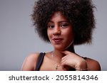 Small photo of Sweet young Black woman with an impish grin and her finger to her chin smiling quietly at the camera in a close up cropped portrait over a grey studio background