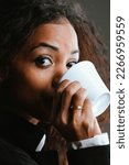 Small photo of Portrait of Beautiful Woman with black skin and long wavy hair, elegantly dressed, drinking coffee from a white cup. She has an amused and intelligent expression, almost intrigued, but sympathetic.