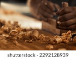 Small photo of The wood artisan, a robust and strong man known as a carpenter, hand planes a board in the traditional manner, producing shavings of sawdust that look like golden curls of wood. It is a strenuous and