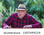 Small photo of An elderly man in a hat is sitting with bad mood at a table in the garden. Concept: evil and unsympathetic people