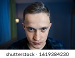 Small photo of Young man with paranoid schizophrenia looks out from under his eyebrows with hatred. An embittered guy with gray eyes and a short haircut against a dark blue room. 22 year old boy with an angry look.