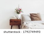 Cup of coffee and books on retro wooden bedside table. Rustic white ceramic vase with bouquet of pink cocmos and zinnia flowers. Beige linen and velvet pillows in bed. Scandinavian interior, bedroom.
