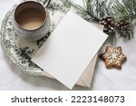 Small photo of Christmas still life. Blank greeting card, invitation mockup.Gingerbread cookie, cup of coffee and pine tree branches. Floral green plate. White linen tablecloth with envelope. Winter festive flatlay.