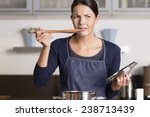 Small photo of Attractive young female cook standing at the hob in her apron tasting her food in the saucepan with a grimace as she finds it distasteful and unpalatable