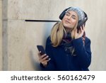 Young blonde woman in coat and hat standing outside against concrete wall holding smart phone in hand with eyes closed smiling listening to music in wireless headphones