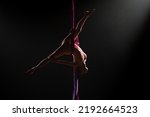 Small photo of Female circus gymnast hanging upside down on aerial silk and demonstrates stretching. Woman performs tricks at height on red silk fabric. Difficult acrobatic stunts on black background with