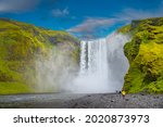 Powerful and famous Skogafoss waterfall with a lonely standing person in orange jacket, while hiking in Iceland, summer, scenic dramatic view at sunny day and blue sky.