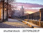 Prague, Czech Republic. Sunrise sunshine sunbeams and shadows on paving stones from promenade from river Vltava at old Winter street with trees and lantern. Tower Charles Bridge. Scenic sky clouds.