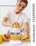 Small photo of Young beautiful woman preparing orange juice with modern citrus juicer machine at home