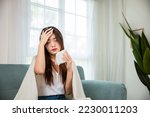 Small photo of Sick woman sitting under blanket on sofa and sneeze with tissue paper in living room, Asian young female blowing nose sneezing in tissue at home, fever caught cold