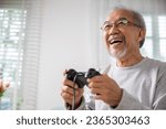 Small photo of Asian senior old man enjoying holding joystick playing video game at home in living room, mature man hands using game controller, Funny on retirement elderly smile sitting on sofa life gaming