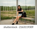 A young beautiful girl in denim shorts and with a backpack sits at a bus stop waiting for a bus and looks at her watch.
