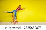 Small photo of A beautiful young woman in a red plaid shirt, blue jeans and white sneakers stands on a red stepladder and in an unusual pose with an outstretched leg stretches to paint the wall