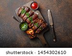 Grilled or fried and sliced marbled meat steak with fork, tomatoes as a side dish and different sauces on wooden cutting board, top view, close-up, stone rustic background. Beef meat steak concept 