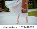 Detail of young fashionable woman wearing beige coat, tulle midi skirt and gold high heels. She holding stylish beige handbag with silver details in hands. Street style.