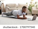 Training At Home. Sporty man doing yoga plank while watching online tutorial on laptop, exercising in living room, free space
